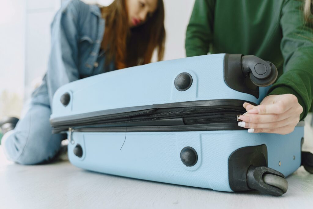 Two people struggling to close a blue suitcase by pulling the zipper